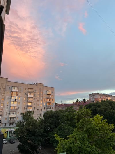 The image depicts a serene evening sky over an urban landscape in Kropyvnytskyi, Ukraine. The sky is painted with soft hues of pink and pale blue, suggesting a sunset or twilight scene. Wispy clouds are scattered across the sky, catching the fading light and adding depth to the celestial canvas. Two thin power lines cut diagonally across the upper left portion of the frame.
Below the sky, several apartment buildings dominate the scene. These multi-story structures, typical of Soviet-era architecture, feature rows of windows and balconies. Some windows are illuminated, hinting at the ongoing daily lives within. The buildings are rendered in shades of gray and beige, with some showing signs of wear. Lush green trees stand tall between the buildings and in the foreground, their dense foliage providing a natural contrast to the concrete structures.
A glimpse of a street can be seen at the bottom right, with a few parked cars, including a dark SUV and a white sedan. A small sign with Cyrillic text is visible on a building façade, likely indicating a business. No people are clearly discernible in the image, giving the scene a quiet and contemplative feel. The overall atmosphere is one of peaceful transition from day to night in a residential area.