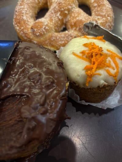 The image shows a close-up of three pastries on a dark surface. From left to right, there is a sugar-dusted pretzel, a carrot cake cupcake with white frosting and shredded carrot topping, and a chocolate-glazed pastry, likely a croissant or similar viennoiserie. A pair of metal tongs is partially visible, positioned near the pretzel and cupcake. The pastries appear to be from a bakery in Mexico City, Mexico.
