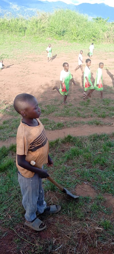 In Ngalula, Democratic Republic of the Congo, a young boy stands on a grassy patch of land holding a hoe. He is wearing a striped brown t-shirt, pinstripe pants, and sandals. In the background, several other children are visible on a dirt field, wearing what appear to be sports uniforms. Behind them is a field with tall grass and trees, and further back are mountains.