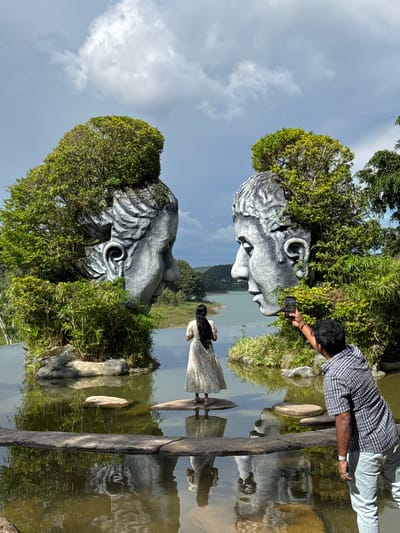 This is a photograph of the Datanla Waterfall area in Da Lat, Vietnam, featuring a prominent art installation of two colossal, stone heads that appear to emerge from the water and foliage. The heads are stylized and weathered, with trees and dense greenery growing from within them, particularly on the tops and sides.
In the foreground, a man wearing a checkered shirt and light-colored jeans is taking a photograph of the scene with his smartphone. He is positioned to the right of the frame, facing the art installation. To the left of the frame, a woman with long, dark hair, wearing a floral-patterned dress, stands on a stone in the water, looking towards the heads. Her reflection is visible in the water.
The art installation is situated on a body of water, possibly a lake or a wide section of the river near the waterfall. The water is calm and reflects the sky, clouds, and the surrounding landscape. The background shows a hilly landscape with trees and a body of water, suggesting the natural beauty of the Da Lat region. The sky is partly cloudy, with patches of blue and large, white clouds. The overall scene is a blend of natural scenery and artistic sculpture, creating a unique and visually striking composition.