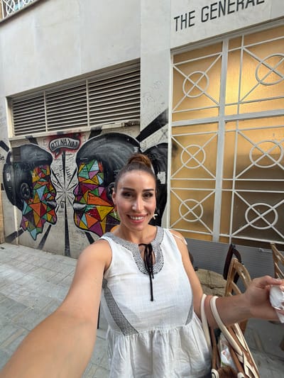 This is a selfie taken outdoors, possibly during the daytime due to the bright lighting. The subject is a woman with her hair pulled up, smiling at the camera. She is wearing a white dress with black trim. In the background, there is a wall adorned with vibrant graffiti depicting two faces composed of geometric shapes and patterns. The word "OTI NA WAI" is visible on the graffiti. To the left of the graffiti, there is a white building with the text "THE GENERAL" written on it in black. Above the graffiti and to the left, a white metal gate with a gold-colored translucent material behind it is visible. To the right of the woman, there are some wooden objects, possibly part of a market stall or display, and a person's arm holding a white tissue. The overall scene suggests an urban environment with artistic elements.