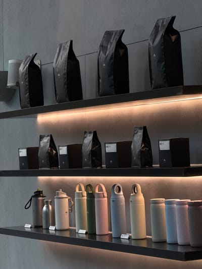 The image depicts a retail display featuring numerous coffee bags and reusable water bottles, arranged on dark shelves in a dimly lit store. On the top shelf, several large, black, matte-finish coffee bags with a white "M" logo are prominently displayed. To the left of these bags, a white cylindrical object is partially visible. The middle shelf holds smaller, dark boxes and black coffee bags, some of which have labels with white text, including "#45 blended". The bottom shelf showcases a collection of vibrantly colored reusable water bottles in various shades of grey, beige, white, green, and pink, each with a white "M" logo or similar marking. The overall aesthetic is modern and minimalist. The scene is consistent with a specialty coffee shop or boutique retail space. The image is described as being located in Xiangzhou, China, suggesting this is a commercial establishment within that region.