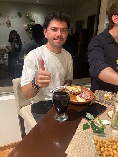 A man in a white long-sleeved shirt is seated at a table with a glass of red wine and a charcuterie board with meats and cheeses. The table is also set with a small appetizer, a smartphone, greenery, and a container of nuts. In the background, there are balloons and a "Happy Birthday" banner. Other people are visible in the background as well. Based on the characteristics of the architecture and the decorations, the location is likely a residence or event space in Mexico City, Mexico.