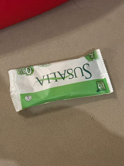 A vertically oriented snack bar, predominantly white and green, rests on a textured tan surface, partially overlapping a red object. The bar features the brand name "SUSALLA" in large, vertically stacked lettering. A "BIO" label is visible at the top, and a "0%" symbol is at the bottom. The wrapper has a crinkled texture and a serrated top edge.