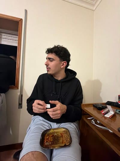 An indoor scene in Bragança, Portugal, features a young adult male seated, angled to his left, with his hands positioned centrally. He has dark, curly hair, a mustache, and a soul patch, and is wearing a black Carhartt hooded sweatshirt and light-colored shorts. His hands are actively engaged in rolling a small white paper. On his lap rests a rectangular, metallic tray containing loose organic plant material. To the right, on a wooden surface, a dark-colored handgun is visible, positioned near a red and white package and several white cables. A wooden-framed mirror on the left wall reflects the back and upper head of another individual, dressed in dark clothing and light grey pants. The room walls are plain and light-colored, featuring simple crown molding at the ceiling.