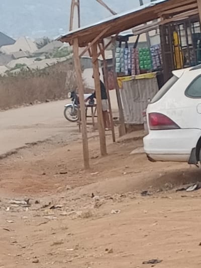 A roadside shop is visible. The shop is a simple wooden structure with shelves stocked with items. A white car is parked partially in front of the shop. A motorcycle is visible near the shop. The area around the shop is a dirt road. The image is taken at a slightly skewed angle.