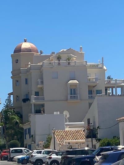 The image shows a multi-story building with a dome on top, located in Estepona, Spain. The building is painted in a light color and has multiple balconies and windows. In front of the building, several cars are parked. The sky is clear and blue. The overall scene appears to be a sunny day.