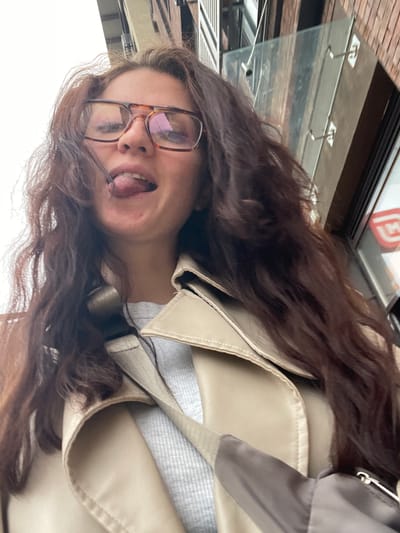A woman with long, brown, wavy hair and glasses is seen in an eye-level, tilted, outdoor shot. She is wearing a tan trench coat and a light grey patterned shirt. Her tongue is sticking out. In the blurred background, there's a building with a glass awning, suggesting an urban environment. The city of Razvilka, Russia is not discernible from the image.