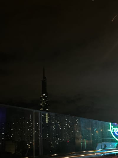 The image captures a nighttime cityscape from a high vantage point, likely a rooftop or a tall building. In the background, a prominent, slender skyscraper dominates the skyline, illuminated by numerous lights, suggesting it is a modern architectural marvel. The sky is dark and overcast, with some faint glows indicating urban light pollution. In the foreground, a glass barrier reflects the city lights, creating a blurred, shimmering effect. The city itself appears densely populated with many buildings adorned with lights, creating a vibrant, twinkling panorama. A neon sign with blue and green lights, partially visible on the right, adds a touch of contemporary advertising to the urban scene. The overall impression is that of a bustling metropolis at night, with the dark sky contrasting against the artificial lights of the city. The presence of the specific neon sign suggests a commercial or entertainment district within Thủ Đức, Vietnam.