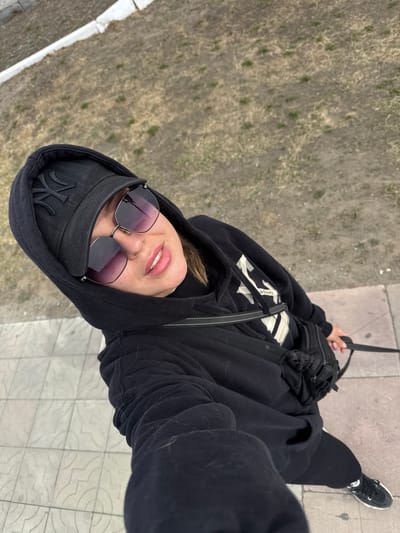 A woman is standing on a stone tiled ledge in Borzya, Russia. She is wearing black pants and a hooded black sweatshirt. She also wears a black baseball cap and sunglasses. She has a black purse on her chest. The ground below is brown dirt with some sparse green vegetation. The edge of a white curb is visible on the left of the frame.