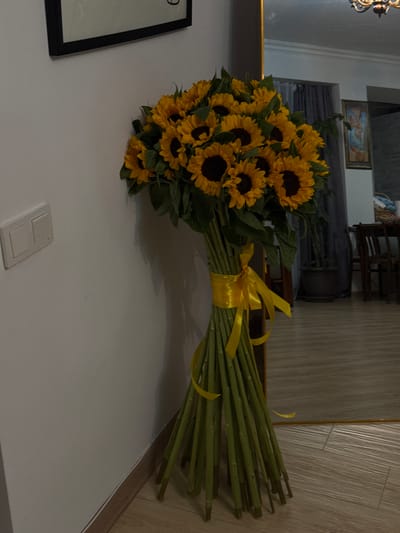 A vibrant, tall bouquet of numerous sunflowers stands upright against a plain white wall in an indoor setting. The sunflowers feature bright yellow petals surrounding dark, prominent centers, and are accompanied by lush green leaves. Their long, green stems are neatly gathered and tied with a sunny yellow satin ribbon that forms a bow, positioned about a third of the way down from the flower heads.

The white wall serves as the immediate background for the bouquet. Below the bouquet, a light-colored floor, possibly made of wood or laminate, is visible, featuring subtle vertical grain patterns. A small, white rectangular electrical fixture, likely a light switch or outlet, is mounted on the wall to the right and slightly below the bouquet's base. On the far bottom left, a black-framed object, possibly a picture, is partially visible, with the number "9" faintly discernible in its top left corner.

To the left and above the bouquet, a large framed mirror reflects elements of the room beyond the immediate scene. Within the mirror's reflection, parts of a dark piece of furniture, possibly a bed or sofa with dark gray upholstery, can be seen. Above this furniture, a small framed painting depicting classical figures in light blue and beige tones hangs on the wall. Further back in the reflection, a dark, intricate wooden structure, resembling part of a chair or decorative screen, is also visible. The reflected floor appears consistent with the visible floor.

The lighting suggests a well-lit indoor environment, possibly a home, though no explicit time of day or weather conditions are indicated. The sunflowers imbue the scene with a cheerful and warm atmosphere. No specific activity is taking place; the bouquet appears to be displayed. There are no visual cues in the image to specifically confirm the location as Yerevan, Armenia.