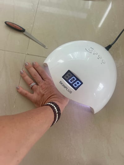 The image shows a woman's hand being held under a white, dome-shaped UV nail lamp on a tiled surface. The lamp displays "8.8.8" on its digital timer and is labeled "SKINPLUS." To the left of the lamp, a metal nail file with a black and brown handle is visible. The woman is wearing a silver ring and beaded bracelets. The location is Puttaparthi, India.