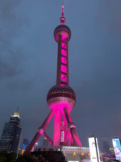 This is a low-angle, full-shot photograph of the Oriental Pearl Tower in Pudong, China, at dusk. The iconic television tower is illuminated with vibrant pink lights, highlighting its spherical structures and supporting legs. In the background, other skyscrapers of Pudong are visible, some lit with their own colorful lights, while the sky is a deep, cloudy blue. The foreground shows the base of the tower with some surrounding structures and trees, and large digital billboards displaying images and text are also present. The overall scene conveys a sense of urban grandeur and nighttime city life.