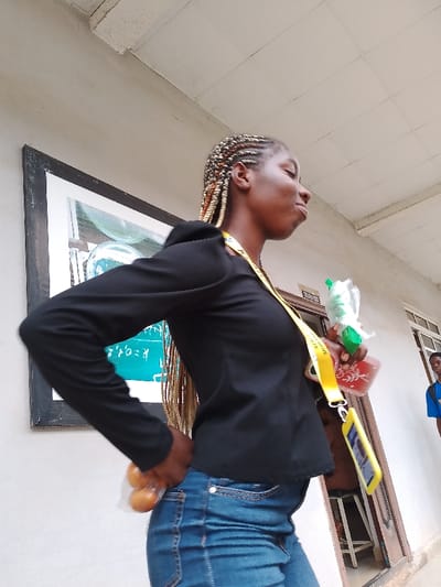 A young, dark-skinned woman stands in front of a white building in what appears to be Umueshi, Nigeria. She is wearing a black long-sleeved shirt and blue jeans. She has braided hair with blonde highlights. A yellow lanyard hangs around her neck, holding a name tag and a cell phone. In her right hand, she holds a clear plastic bag with what appears to be a green fruit or vegetable, as well as a red container. Her left hand is placed in her jean pocket. To the left of her is a framed picture on the wall. Behind her, a doorway and a partial view of a young man in a blue shirt are visible. The setting seems to be outdoors with natural lighting.
