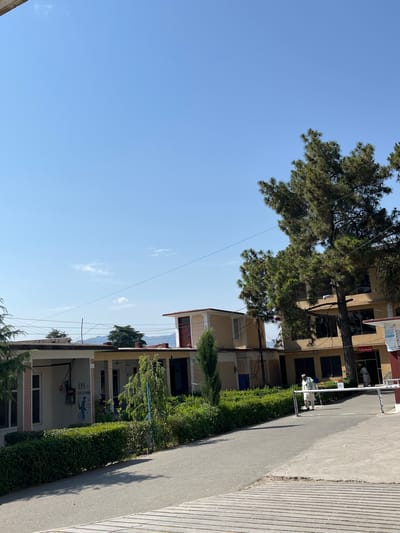 This image captures an outdoor scene at the EPI Center in Kabal, Pakistan, under a clear blue sky with sparse, wispy clouds. Distant mountains are visible in the background, partially obscured by intervening structures and trees.
The foreground features a paved asphalt road that curves towards a complex of buildings. The immediate foreground shows a textured, ribbed concrete or wooden surface. The road is bordered on the left by a neatly trimmed green hedge.
On the left side of the scene, a light-colored, single-story building with a covered porch is visible. A sign affixed to its wall clearly reads "EPI CENTER KABAL" in English, positioned above a cartoon-like mural of a blue character holding a syringe. A grey utility box is mounted on the wall.
Towards the center-right, a taller, multi-story building with a light brown or beige facade features several dark windows and a prominent red entrance door. Adjacent to this door is a white sign with red text, largely in Urdu, which translates to "Registration Ward." Large, mature coniferous trees with dark green foliage partially obstruct views of this building.
Overhead, multiple power lines crisscross the scene. A metal barrier or gate structure is present on the right side of the road.
Two individuals are visible: one male, dressed in traditional white shalwar kameez and a white head covering, is walking leftward on the paved path near the metal barrier. Another figure in light-colored attire is partially visible within the doorway of the multi-story building.