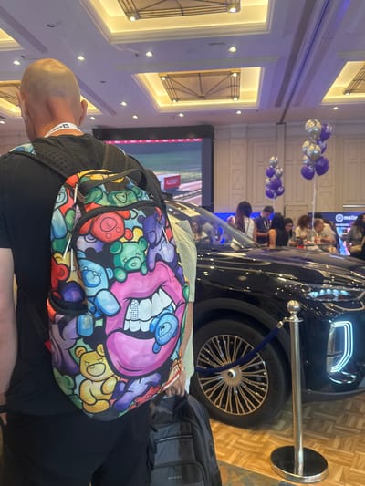The image shows a man with a bald head wearing a black t-shirt and black pants, carrying a backpack with a colorful design featuring teddy bears and lips. He is standing in an indoor venue, possibly a convention center or exhibition hall, with a large screen displaying an image in the background. A black car is parked behind him, and a crowd of people are visible in the distance. Balloons are also present, some of which appear to be branded. The floor is covered in a wooden parquet pattern. There is no indication that the location is in Tel-Aviv, Israel.