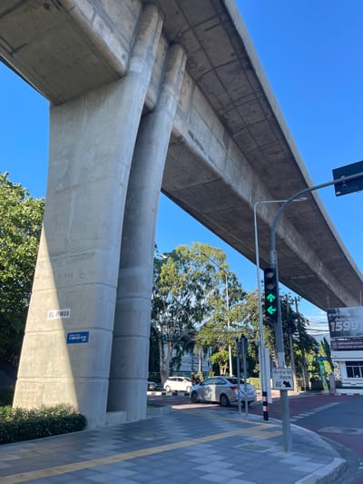An overpass for an elevated transit system, likely the BTS Skytrain or MRT, arches across a clear blue sky in Bangkok, Thailand. The massive concrete structure is supported by substantial pillars, one of which bears a label reading "BL-PW08" and a blue sticker with a phone number. Beneath the overpass, a traffic light displays green arrows, indicating traffic flow. In the foreground, a pedestrian walkway with tactile paving leads towards a street intersection. Several vehicles, including cars and a motorcycle, are visible on the road. Lush green trees frame the scene, and a billboard advertising a service is positioned on the right. The overall impression is one of urban infrastructure integrated into the city's environment.