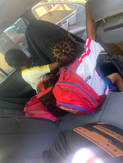 The image depicts the interior of a vehicle, possibly a car, with three individuals visible. Two children are seated in the back, and an adult appears to be in the front. The children have backpacks. The environment appears to be a street in Sabon Tasha, Nigeria. The image is taken from a slightly elevated angle looking down into the car.