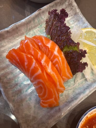 A close-up, overhead shot features three pieces of raw salmon sashimi arranged on a light green and blue speckled rectangular plate. The salmon is a vibrant orange with thin white fat lines. To the right of the salmon, there is a small pile of shredded daikon radish, a sprig of dark red lettuce, and a slice of lemon. The plate sits on a dark, textured surface, likely a table. In the lower right corner, a small white dish with a blue rim contains a red sauce. The scene appears to be from a restaurant in Kuta, Indonesia, given the context provided.