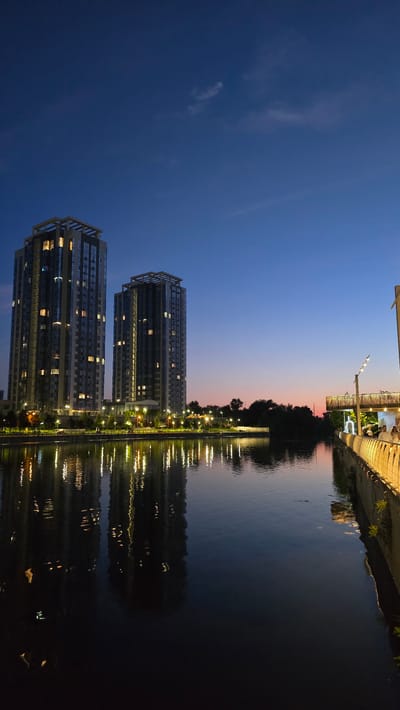 The image depicts a twilight scene in Kyiv, Ukraine, likely along the Dnipro River or one of its tributaries. In the background, two modern high-rise apartment buildings stand illuminated, with lights visible in many of their windows. The sky is a gradient of deep blue at the top, transitioning to hues of pink and orange near the horizon, indicating the end of daylight. A body of water occupies the foreground and midground, reflecting the lights of the buildings and the sky. Along the right bank of the water, a pedestrian walkway or promenade is visible, featuring decorative lighting and a railing. A few indistinct figures can be seen walking on this promenade. Trees and greenery are present along the banks, separating the buildings from the water. The overall atmosphere is serene and urban.