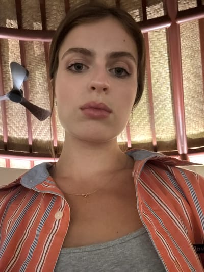 A young woman is captured in a close-up, front-facing shot in Tegallalang, Indonesia. She is wearing a grey tank top under an open, orange and white striped button-down shirt with blue pinstripes. A delicate gold necklace with a knot pendant rests on her collarbone. Her face is the primary focus, with her brown hair framing her face and dark brown eyes looking directly at the camera. Her lips are slightly parted, and her skin has a natural glow. Behind her, a woven bamboo wall forms the backdrop, with vertical wooden beams painted in a reddish-brown hue. A portion of a ceiling fan is visible in the upper left corner of the frame, its dark blades contrasting with the lighter background. The lighting appears to be natural and soft, suggesting an indoor setting with diffused light.