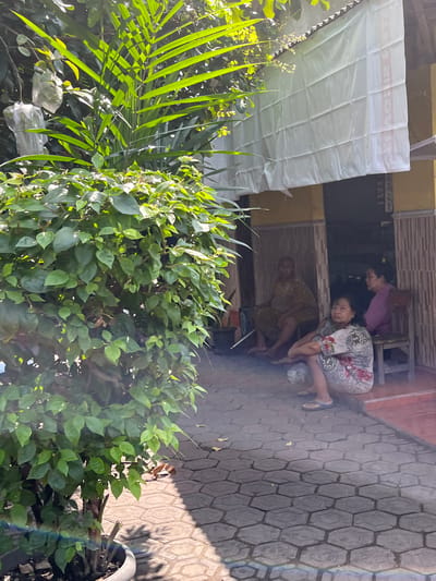 The image depicts an outdoor scene in Jati Alun Alun, East Java, where three individuals are seated on a tiled patio in front of a building with a yellow facade. A large, leafy plant is prominent in the foreground, with its branches casting shadows on the ground. A white sheet is hung above the building's entrance.

In the middle ground, three individuals are seated. The person furthest back, on the left, is wearing a light brown garment and is sitting on a chair. To their right, a person in a purple top is seated on a wooden chair. In the foreground, closer to the viewer, a person is seated on the ground, wearing a patterned dress. They are facing the camera, with their legs crossed. A plastic bag is visible next to them on the ground. The patio is paved with hexagonal tiles, and some scattered leaves are visible. Sunlight is casting strong shadows, suggesting it is daytime. The overall atmosphere appears relaxed and informal.