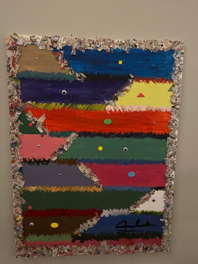 A painting hangs on a light-grey wall. The painting is multi-colored with vertical stripes of blue, yellow, red, green, pink, grey, and brown. The edges of the painting are decorated with pieces of torn paper. Each colored stripe contains small geometric shapes and googly eyes in varying sizes and colors. A dark-blue stylized symbol or signature is present on the right side of the painting.

There is no information about the location of the painting.