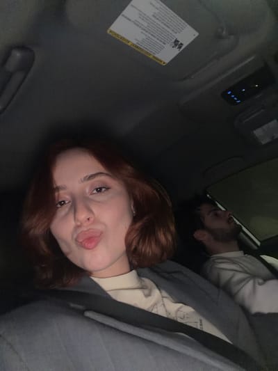 The photograph, apparently taken inside a vehicle, shows a woman with auburn hair sticking out her tongue toward the camera. She appears to be in the passenger seat. A male figure is visible in the background, seemingly seated behind her. The environment is dark, suggesting it is night or a poorly lit space. A warning sticker is affixed to the vehicle’s interior, and various buttons and lights are present on the roof console. There is no visual evidence within the image to determine the location as Grigalati, Georgia, or any specific geographic locale.
