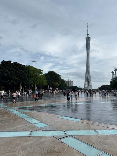 The image shows a wide, wet, paved plaza in front of the Canton Tower, located in the Tianhe District of Guangzhou, China. The sky is overcast with gray clouds. Numerous people are walking across the plaza, some carrying umbrellas, suggesting it has recently rained or is still drizzling. The ground is reflective due to the moisture. In the foreground, the plaza features geometric patterns of stone paving with embedded strips of frosted blue glass or light. The Canton Tower, a distinctive lattice-like observation tower, dominates the background, towering over the scene. Trees line the sides of the plaza, and several other modern buildings and structures are visible in the distance.