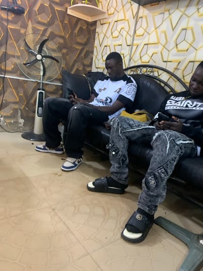 Two young Black men are seated on a black leather sofa in an indoor room. The man on the left is wearing a white t-shirt with black sleeves and graphics, black pants, and blue and white sneakers. He is holding a cell phone in his hands. The man on the right is wearing a black t-shirt with graphics and distressed, dark wash cargo pants, along with black socks and black slide sandals. He is also holding a cell phone. Behind them, the wall features a geometric pattern with gold accents. To the left of the sofa, a white pedestal fan stands. The floor is tiled in a light tan color with a circular pattern. The scene appears to be a casual indoor setting, possibly a lounge or waiting area.