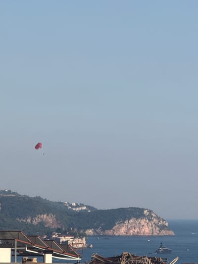 A parasailor with a red parachute is suspended in a clear, light blue sky over the Adriatic Sea. Below, a lush, green, and rocky hillside dotted with white buildings slopes down to the sea. The hillside is part of the coastal landscape near Boreti, Montenegro. In the foreground, a partially visible structure with a metal frame and solar panels suggests a rooftop or balcony area, possibly a hotel or residential building. A large, dark yacht is anchored in the water near the hillside. The water is a deep blue, reflecting the clear sky. The overall scene depicts a sunny day at a coastal resort.