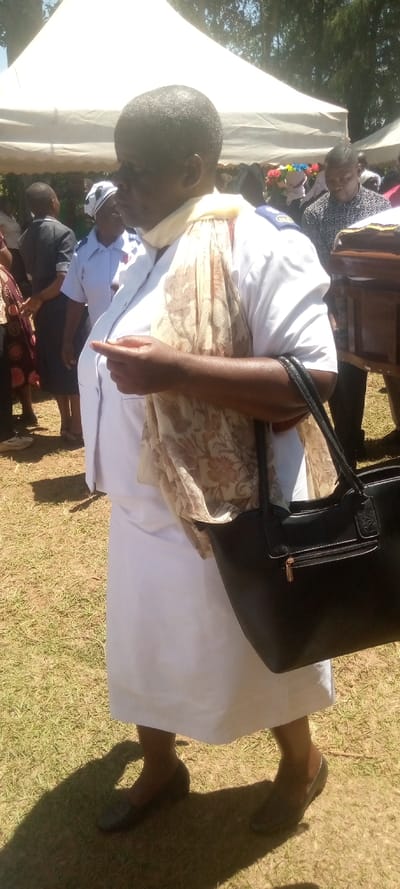 The image shows a woman, wearing a white tunic and a beige patterned scarf, standing in a grassy area. She is holding a black handbag and has black shoes on. The image was taken in Lwandeti, Kenya, at coordinates latitude: 0.537, longitude: 34.799. Behind the woman is a white tent, partially obscured by trees in the background. There is a group of people in the background. There appears to be a wooden casket on a cart to the right of the woman. The woman is looking down at her hand and appears to be adjusting something on her clothing. The environment is outdoors, with grass and trees in the background.