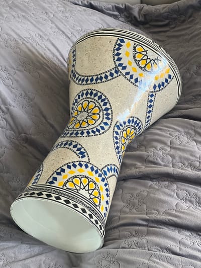 A single doumbek lies horizontally on a gray, quilted blanket. The doumbek is light-colored with yellow, blue, and black geometric patterns. Its wide end faces the lower right of the frame. The surrounding blanket is slightly wrinkled. The image was most likely taken indoors in Armenia.