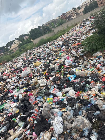 This media file captures a vast expanse of illegally dumped waste, predominantly plastic bags and packaging, piled high along a roadside in Nkpor, Nigeria. The scene is overwhelming, showcasing the sheer volume of refuse that has accumulated. Amongst the debris, a variety of objects are discernible: white and black plastic bags, colorful wrappers, bottles, and discarded containers. The garbage is densely packed, creating an uneven and chaotic landscape.
To the left of the dump, buildings and trees are visible, suggesting an urban or semi-urban setting. The presence of these structures provides a stark contrast to the environmental degradation. The image appears to have been taken during the daytime, with diffused light suggesting it could be overcast or late afternoon. The sky, visible in some crops, is cloudy.
There is no discernible activity or event taking place beyond the existence of the dump itself, implying a static, long-term problem. The overall impression is one of neglect and environmental crisis. Some faint text is visible on packaging, such as "PURITY" on a blue and white packet, and "ipo" on a red and white packet. However, much of the text is obscured or illegible due to the nature of the waste. The scale of the dump and the surrounding environment hint at the significant challenges faced by communities in managing waste in this location.