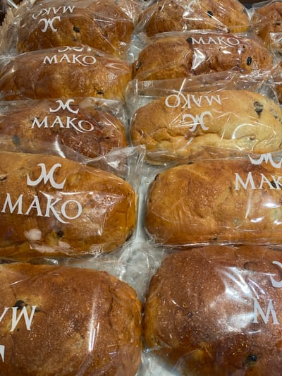 A close-up, high-angle shot shows several loaves of raisin bread in plastic packaging. The bread is golden brown and appears to be fresh. The loaves are arranged in rows, with some visible in the foreground and others behind them. Each loaf has a white logo printed on the packaging that reads "MAKO" with a stylized "M" above it. The background is blurred, but it appears to be a display case or shelf. The lighting is bright and even, suggesting an indoor setting like a bakery or supermarket. There are no people or other animals visible in the image. The scene is likely located in a bakery or food retail establishment in North Jakarta, Indonesia, given the branding of the bread.