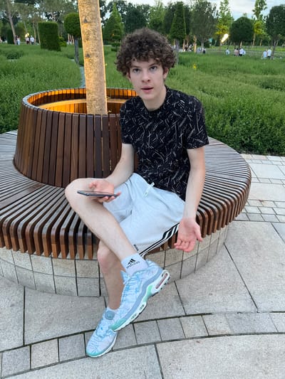 A young male with curly brown hair sits on a curved wooden bench in a park. He is wearing a black t-shirt with a white abstract pattern, white shorts, white socks with black stripes, and light blue and white sneakers. He is holding a black smartphone in his right hand and looking towards the camera with a surprised expression. The bench is located in the city of Krasnodar, Russia, and is surrounded by lush green grass, trees, and a stone pathway. In the background, other people can be seen walking and enjoying the park. The lighting suggests it is late afternoon or early evening.