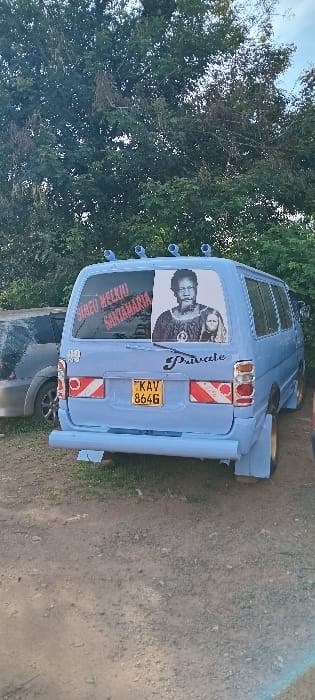 The image displays the rear-left quarter of a light blue van, likely a public transport vehicle (matatu), parked on unpaved ground in Kisumu, Kenya. The vehicle carries a Kenyan license plate, KAV 864G, positioned centrally on the rear. The rear windshield features two distinct visual elements: a diagonal red text sticker on the left, partially legible as "SIMZO MELKIO SANTAMARIA," and a black and white image on the right portraying a bearded man with a younger individual resting on his shoulder. Below the right rear window, the word "Private" is written in a black cursive font. Four small, light blue, cylindrical objects are affixed to the rear edge of the van's roof. Red and white striped reflective tape is applied horizontally across the rear, above the license plate, and on the lower left bumper. The rear wheel wells are fitted with extended, light blue mudguards. To the immediate left of the van, the rear section of a silver SUV is partially visible. The immediate ground is composed of dirt and gravel with sparse green patches, while the background consists of dense green foliage from large trees under a partly visible sky.