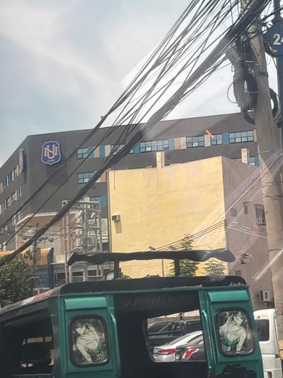 The image shows a street scene in Davao City, Philippines, with a prominent building with a dark facade and a large blue and gold logo that reads "NU 1999." Adjacent to this building is a smaller, yellow building. A green motorized tricycle is in the foreground, with anime-style decals on its windows. Overhead, a complex network of electrical wires crisscrosses the sky, suspended from a utility pole on the right side of the frame, which has a blue sign with the number "24" on it. The lighting suggests it is daytime, with a slightly overcast sky. The background includes other vehicles and some trees.