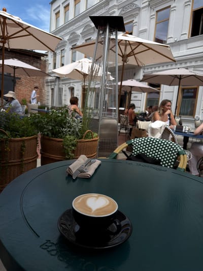 The media file captures an outdoor seating area of a cafe on a sunny day. The scene is dominated by a dark green circular table in the foreground, on which a black cup of coffee with latte art is placed. A napkin with cutlery is also on the table. In the background, several people are visible, some seated at tables, suggesting a relaxed atmosphere. Umbrellas provide shade over some of the seating areas. The architecture of the buildings in the background, with their windows and decorative elements, indicates an urban setting. The overall mood appears to be leisurely and social. The visible text on the table reads "La Bottega".