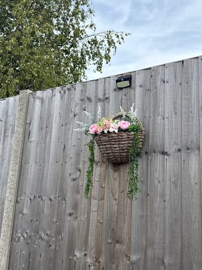 A weathered wooden fence forms the backdrop for a hanging basket filled with artificial flowers and trailing greenery. The basket, made of woven material, is attached to the fence with visible fixings. Above the fence, the branches of a tree laden with green leaves are visible against a cloudy sky. A solar-powered light fixture is mounted on the fence to the left of the basket. A concrete fence post is partially visible on the far left of the image. The scene appears to be in a garden or outdoor area, likely a residential setting in Great Yarmouth, United Kingdom.