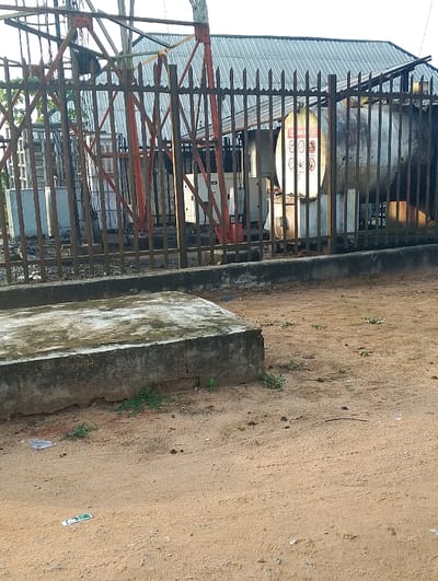 The image shows a fenced-in area with a large, cylindrical tank and a metal structure behind it. There are various warning signs on the side of the tank. There is a large concrete block on the ground in the foreground, with visible signs of moss and cracks. The ground is mostly dirt with sparse patches of grass. The image is taken from a low angle and shows the area through a gap in the fence. The image was likely taken in Ogharefe, Nigeria, as the latitude and longitude provided are located in this area. There are no people or other animals visible in the image.
