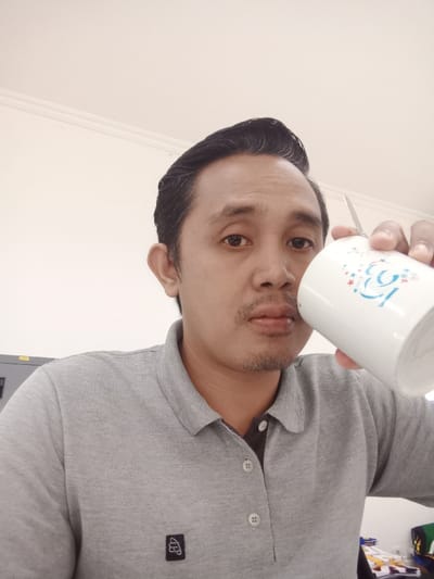 A middle-aged man poses indoors, holding a white mug with a blue design. He wears a gray polo shirt with a small black logo. His hair is dark and neatly styled. The background features a white wall and part of a metal cabinet. The man appears to be looking directly at the camera.