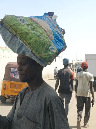 The scene depicts a daylight street view in Fagge C, Nigeria. In the foreground, an adult male, with dark skin and a neutral expression, is seen in left profile. He wears a grey long-sleeved garment with a small chest pocket and has a light-colored band around his forehead. He carries a large, irregularly shaped bundle on his head, consisting of a green and blue woven polypropylene sack printed with "NET WT: 25KG" and additional text including "NIGERIAN".
Behind the foreground subject and to his left, a yellow-orange auto-rickshaw (tricycle) is parked. Its side bears commercial lettering, including "GTMobile", "GAS/KER/GAS", "SUPER", "GAGAICHI", and "GT DATAP".
Further down the unpaved street, three male figures are walking away from the camera. The closest wears a dark t-shirt with orange accents and a blue cap. Beside him, another wears a light-colored t-shirt, dark pants, and a red cap. Further beyond them, a male is seen riding a motorcycle. The background features light-colored walls, indicating buildings, and extends into a bright, clear sky. A white commercial vehicle, possibly a truck or van, is partially visible on the far right in the distant background.