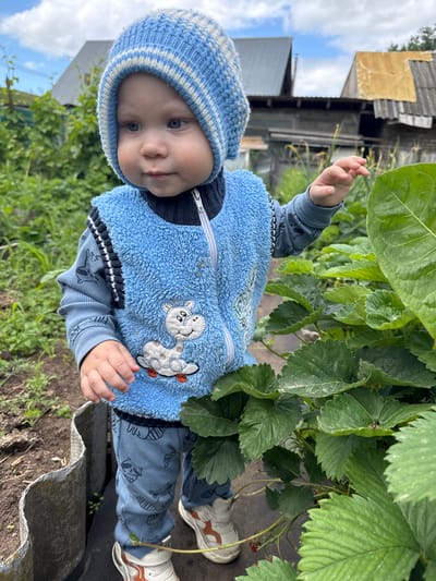 A young child, likely a toddler, stands in a garden wearing a blue knitted hat and a fuzzy blue vest with a cartoon giraffe applique. The child is also wearing blue pants with a raccoon print and white sneakers with orange accents. The child's right hand is extended towards a large green leaf. In the background, there are wooden buildings with corrugated roofs and lush green foliage. The ground appears to be soil and covered with dark fabric. The sky is partly cloudy. The location is identified as Olkhovkа, Russia.
