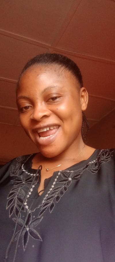 The image displays a close-up, indoor portrait of a young adult female, likely of African descent. She is smiling broadly, revealing her upper teeth and part of her tongue, with her gaze directed towards the camera. Her dark hair is pulled back, and her skin appears dark brown. She is wearing a dark-colored top with intricate white and black embroidery around the V-neckline. A thin, gold-colored necklace with a small spherical pendant rests on her collarbone. The background is a plain reddish-brown surface, possibly a wall or ceiling, with two faint parallel lines visible in the upper portion, indicating an indoor setting. The specific location within the city of Adankolo, Nigeria, cannot be determined from the visual content.