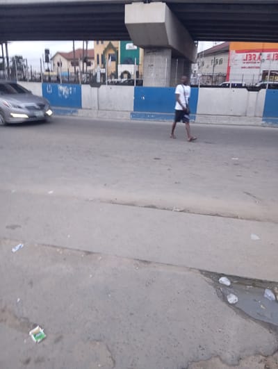 The image shows a street scene in Port Harcourt, Nigeria, possibly under an overpass. A silver car is driving on the left side of the road. A man is walking across the road from right to left. He is wearing a white t-shirt, dark shorts, and sandals, and carrying a bag slung over his shoulder. In the background, there are buildings and a large concrete structure that appears to be part of an elevated road or bridge. The road surface is asphalt and appears worn, with some litter visible, including a crumpled paper packet and some plastic wrappers. The sky is overcast.