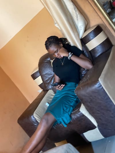 An individual is reclined on a couch in an interior setting, seemingly in Unguwar Gabas, Nigeria. The person has dark skin, braided hair, and is wearing a black top and a teal skirt. They are holding a dark-colored object, possibly a phone. The couch is upholstered in brown and beige leather. The walls are painted a tan color. There is an additional couch or furniture piece partially visible in the background. The image appears to be taken indoors, during daylight. The individual is relaxed and possibly using their phone.
