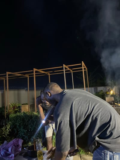 This image captures a nighttime scene in what appears to be a residential outdoor area, possibly a backyard or compound, in Bar Jirgi Summa, Nigeria. The foreground is dominated by the back of a person wearing a dark gray t-shirt and blue jeans, their torso bent as if tending to something. To their left, another person is visible, wearing a dark tank top and light-colored shorts. This second person is illuminated by a bright light source held in their hand, possibly a phone or flashlight, directed downwards. In front of them, a black pot and a clear plastic bottle filled with yellow liquid, likely oil, are placed on a surface, suggesting food preparation or grilling. A crumpled pink plastic bag is also present. In the background, a wooden structure resembling a trellis or pergola is visible, and beyond that, a wall and dense green foliage. Smoke drifts upwards into the dark night sky, indicating a fire or cooking activity. The overall atmosphere is casual and social, with a focus on an outdoor gathering or meal preparation.