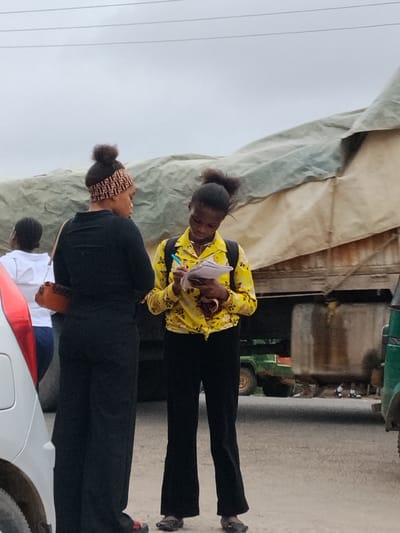 Two young women stand on a street in Aba, Nigeria. One woman wears black pants, a black top, and a Fendi headband, holding a small brown purse. The other wears black pants and a yellow floral shirt, writing on a piece of paper with a pen. Behind them is a large truck covered with a tan tarp, and parts of vehicles are visible on both sides. In the background, other people can be seen near the truck. The sky is overcast. The road surface appears to be paved.
