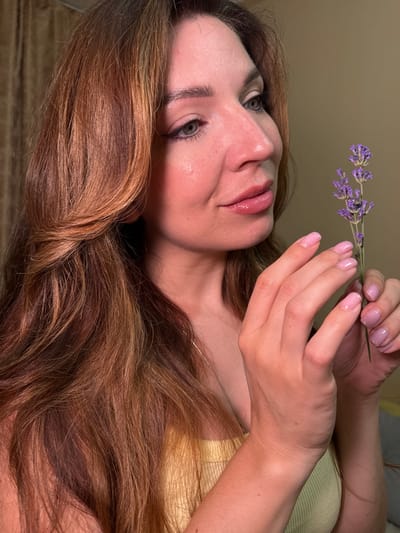 The image features a close-up, vertically oriented shot of a woman holding a sprig of lavender. The woman's face is partially visible, tilted upwards and to the left, with her eyes looking slightly upwards and to the left as well. Her lips are glossy and slightly parted. Her hair is long, wavy, and a warm brown color with lighter highlights, cascading down and obscuring parts of the background.
Her hands, with neatly manicured pink nails, are positioned near her face, gently holding the lavender sprig. The lavender is a delicate cluster of small purple flowers. The background is a muted, warm brown, and a hint of a pale yellow garment is visible on her shoulder. The lighting appears soft, suggesting an indoor setting, possibly during the daytime with diffused light. There are no discernible environmental cues to indicate a specific location within Odesa, Ukraine, nor is any text visible in the image. The overall mood is serene and contemplative.